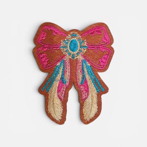 May include: Embroidered patch in the shape of a bow. The bow is brown with pink and blue accents. The center of the bow features a blue jewel. The patch has a decorative, ornate design.