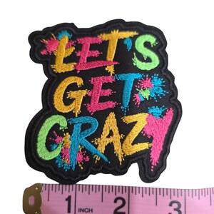 May include: An embroidered patch with a black background and the text "LET'S GET CRAZY" in multi-colored, paint-splatter style lettering. A pink measuring tape is positioned below the patch, showing it is approximately 3 inches wide.
