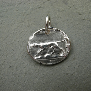 May include: Silver charm featuring a detailed, three-dimensional design of a running dog. The charm has a circular shape and a textured surface.