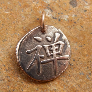 May include: A round copper pendant with the Japanese character for "Zen" inscribed on it. The pendant is on a brown textured surface.