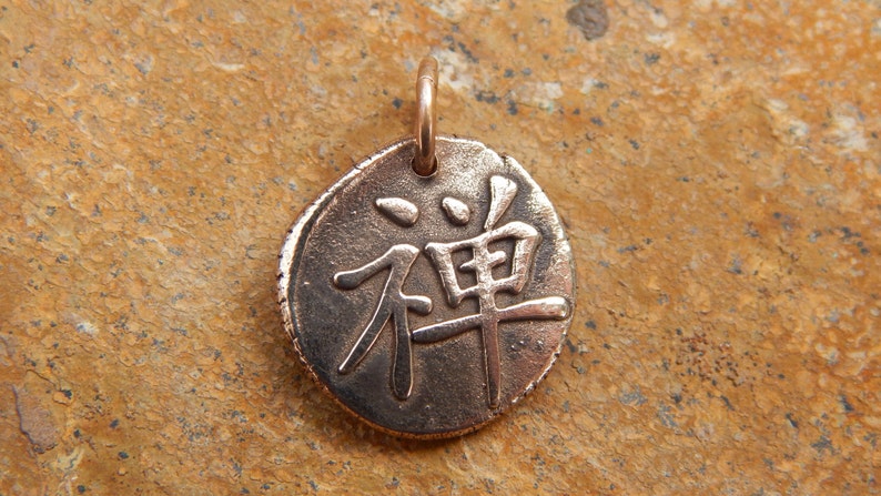 May include: A round copper pendant with the Japanese character for "Zen" inscribed on it. The pendant is on a brown textured surface.