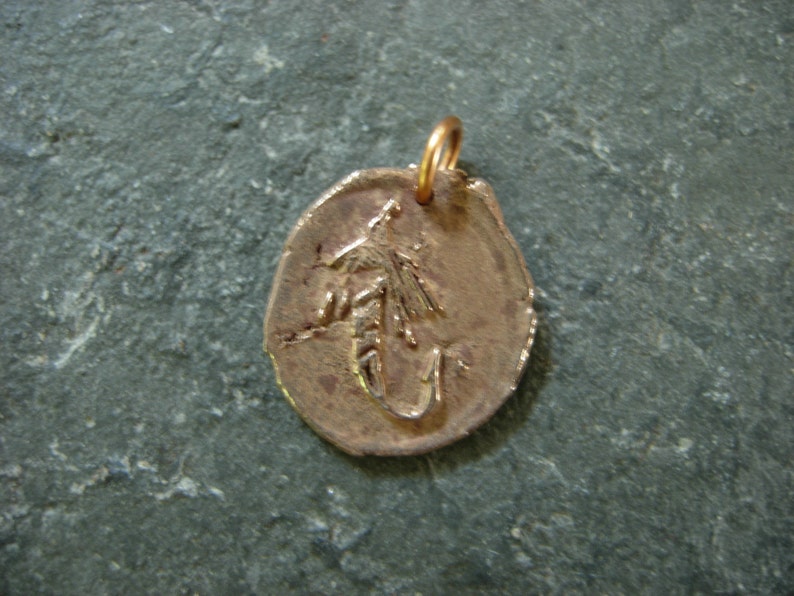 May include: A bronze-colored pendant with a detailed fly fishing lure design. The pendant has a small loop at the top for a chain. The pendant is approximately 1 inch in diameter.