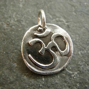 May include: A silver-colored pendant featuring the Om symbol. The pendant has a circular shape with a raised Om symbol in the center. The pendant has a loop at the top for a chain. The background is a dark, textured surface.