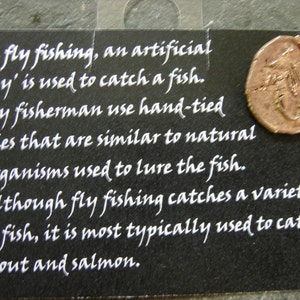 May include: Black card with white text about fly fishing, including the use of artificial flies to catch fish. A copper-colored circular pendant with a fishing fly design hangs from the top.
