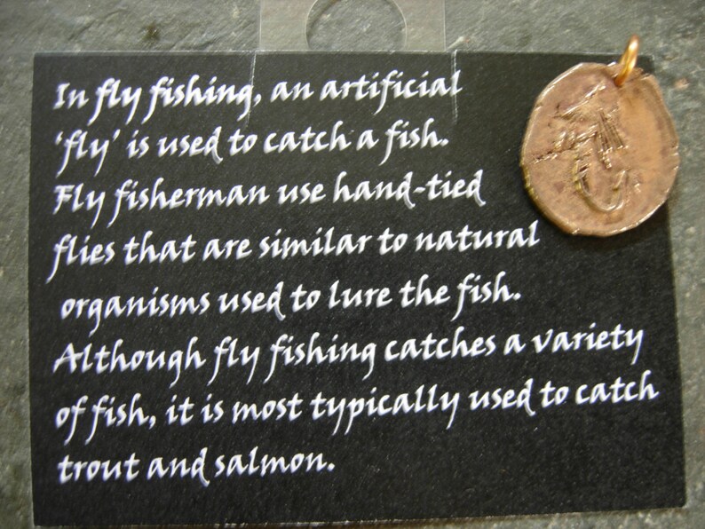 May include: Black card with white text about fly fishing, including the use of artificial flies to catch fish. A copper-colored circular pendant with a fishing fly design hangs from the top.