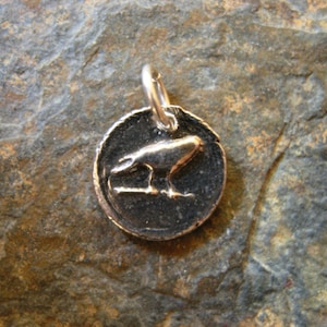 May include: A silver charm in the shape of a round disc with a raised image of a bird. The bird is facing left and has its wings spread out. The charm is on a dark gray background.