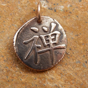 May include: A round, bronze pendant with the Japanese word "Zen" engraved on it. The pendant has a loop at the top for attaching to a chain or cord.