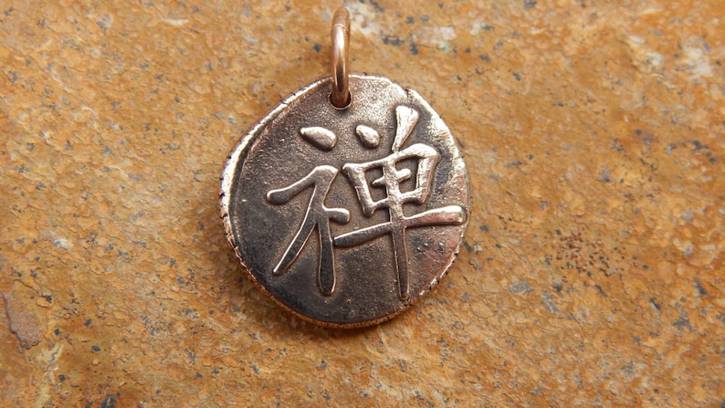 May include: A round, bronze pendant with the Japanese word "Zen" engraved on it. The pendant has a loop at the top for attaching to a chain or cord.