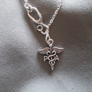 Può includere: Collana color argento con un ciondolo a forma di stetoscopio e un simbolo medico con le lettere "POA". Il ciondolo pende da una delicata catena, su uno sfondo di tessuto grigio. Un accessorio a tema medico.