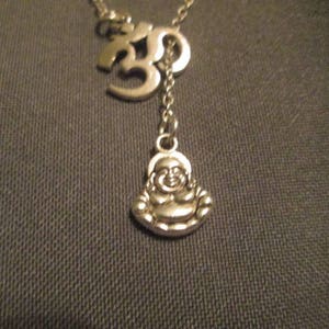 May include: Silver-toned necklace featuring an Om symbol and a small Buddha pendant. The necklace has a delicate chain and a Y-shaped design. The Buddha pendant is smiling. The necklace is likely made of metal.