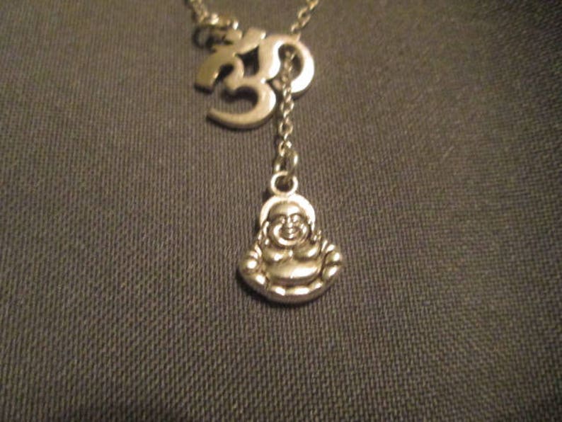 May include: Silver-toned necklace featuring an Om symbol and a small Buddha pendant. The necklace has a delicate chain and a Y-shaped design. The Buddha pendant is smiling. The necklace is likely made of metal.