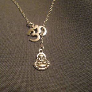 Puede incluir: Collar plateado con un símbolo Om y un pequeño colgante de Buda. El collar tiene una cadena delicada y un diseño en forma de Y. El colgante de Buda está sonriendo. El fondo es una tela gris oscuro.