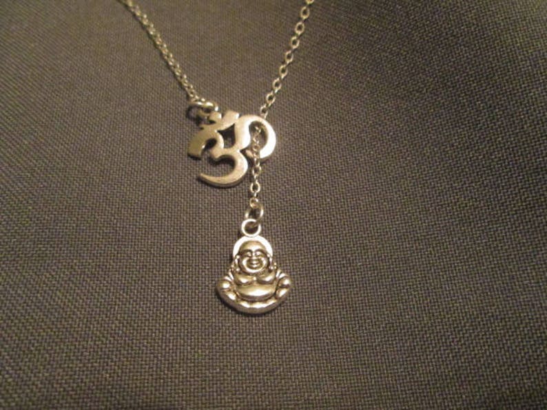 May include: Silver-toned necklace featuring an Om symbol and a small Buddha pendant. The necklace has a delicate chain and a Y-shaped design. The Buddha pendant is smiling. The background is a dark gray fabric.