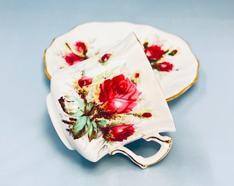 May include: A white porcelain teacup and matching saucer set featuring a vibrant red rose floral pattern with green leaves and gold trim along the scalloped edges. The teacup has a curved handle and the saucer has a matching floral design.