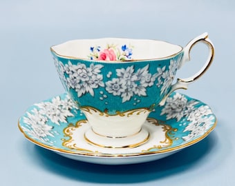 May include: A vintage teacup and saucer set. The teacup and saucer are teal with white floral patterns. The cup has a gold handle and trim. The saucer has a gold trim. The teacup has a white interior with pink and blue floral accents.