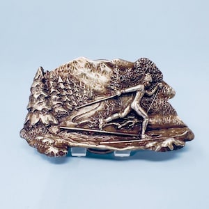 May include: A silver-coloured metal belt buckle depicting a skier. The buckle has a detailed relief design of a skier in a snowy landscape with trees and mountains. The buckle is rectangular with a classic clasp.