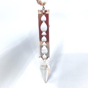 May include: A clear crystal pendant with a rectangular body and a pointed bottom. The pendant features a series of oval and teardrop-shaped cutouts along its length. A small crystal bead is at the top, connected by a metal loop.