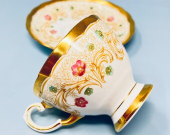 May include: White teacup and saucer set with gold trim and floral accents. The teacup features a curved handle and is decorated with red, green, and gold floral patterns. The saucer has a matching design, creating an elegant tea set.