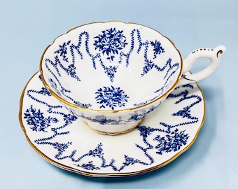 May include: White teacup and saucer set with blue floral patterns and gold trim. The teacup has a scalloped rim and a delicate handle. The saucer also features the same blue floral design and gold trim. The set is placed on a light blue surface.