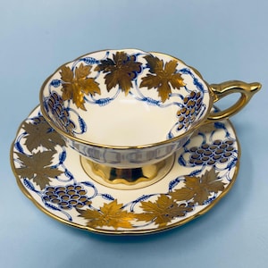 May include: A vintage teacup and saucer set. The cup and saucer are white with gold trim and feature a design of gold leaves and blue grapes. The handle and base of the cup are gold. The set is on a light blue background.