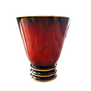 May include: A conical vessel featuring a deep red and black gradient finish with a smooth, glossy texture. The base is composed of three stacked, dark circular tiers, each accented with a thin, metallic gold rim along the bottom edge.