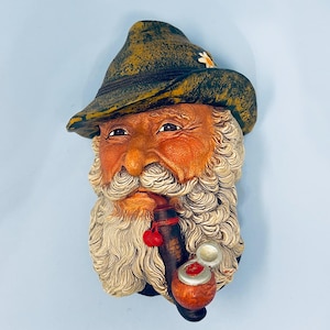 May include: A detailed, hand-painted wall sculpture of an elderly man with a long, white beard and a traditional green hat. The man is smoking a pipe with a red cherry-shaped decoration. The sculpture has a rustic, aged appearance.