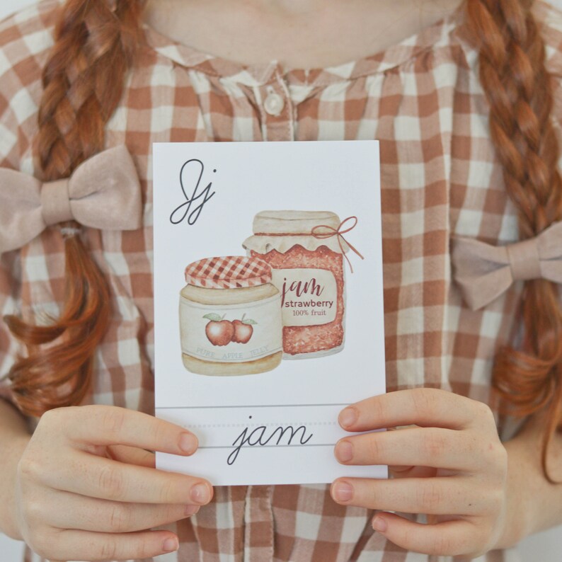 Pu&ograve; includere: Un cartoncino bianco con la lettera 'J' scritta in corsivo in alto. Sono raffigurati due vasetti di marmellata, uno con un coperchio rosso e l'altro con un coperchio bianco. Il testo sui vasetti recita "jam strawberry 100% fruit" e "home apple jelly". La parola "jam" &egrave; scritta in corsivo in basso sul cartoncino.