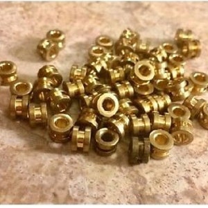 May include: A large group of gold-colored metal beads, each with a raised ring around the center. The beads are arranged in a random pattern on a light brown surface.