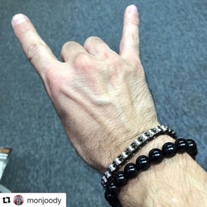 Può includere: Una mano che fa il gesto delle corna, con due braccialetti. Un braccialetto è fatto di perline nere, l'altro è argentato con un design a catena. Lo sfondo è di colore grigio.
