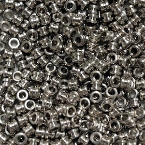 May include: A large pile of silver metal eyelets. The eyelets are small, round, and have a hole in the center. They are used for attaching fabric or other materials together.