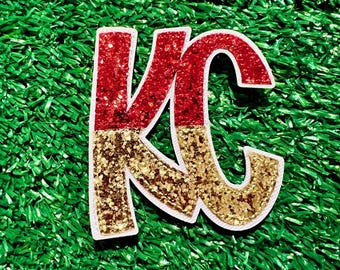 May include: Red and gold glitter "KC" letters on a green artificial grass background.