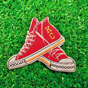 May include: Red embroidered patch of two high-top sneakers with white laces and a yellow stripe. The sneakers are crossed over each other and the top sneaker has the letters "KC" embroidered in yellow.