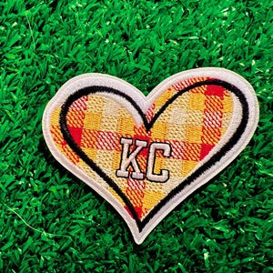 May include: A red, yellow, and white plaid heart-shaped patch with a white outline and the letters "KC" in white inside. The patch is on a green artificial grass background.