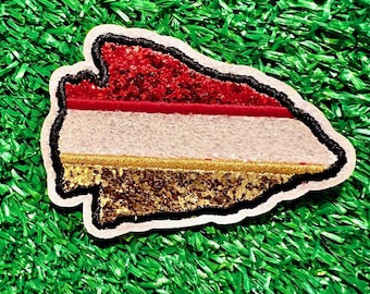 May include: A red, white, and gold arrow-shaped patch with a glittery finish. The patch is on a green artificial grass background.