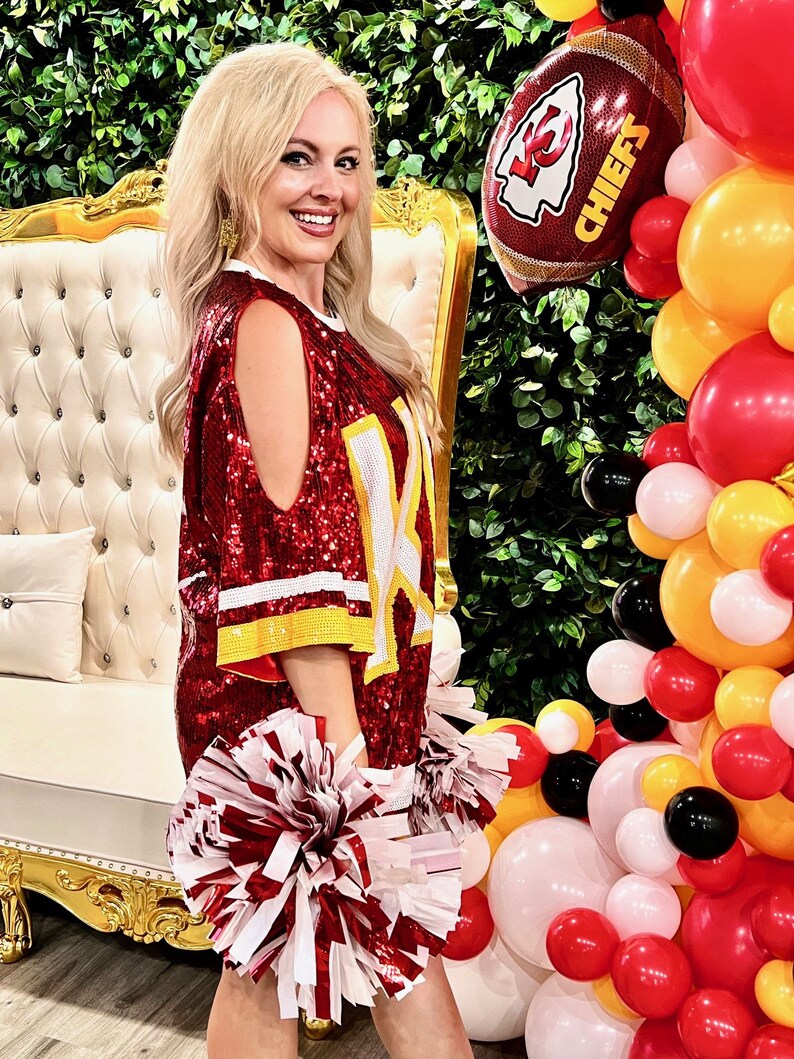 May include: A woman wearing a red sequined jersey with the word "CHIEFS" on it, holding red and white pom-poms. She is standing in front of a white chair and a balloon arch with red, yellow, and black balloons.