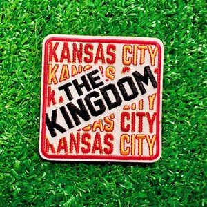 May include: A white patch with red and orange stitching. The patch features the text "KANSAS CITY" repeated multiple times in a diagonal pattern. The text "THE KINGDOM" is in black and centered on the patch.