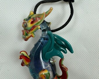 May include: A glass dragon pendant with a blue body, green wings, and red, yellow, and green swirls. The dragon is attached to a black cord.