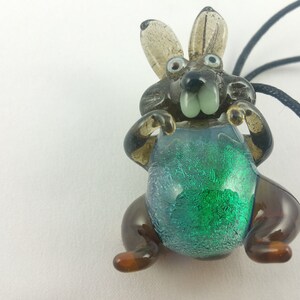 May include: A glass rabbit pendant with a green and blue iridescent body, brown legs, and black eyes. The rabbit is wearing a blue and green iridescent bib. The pendant is attached to a black cord.
