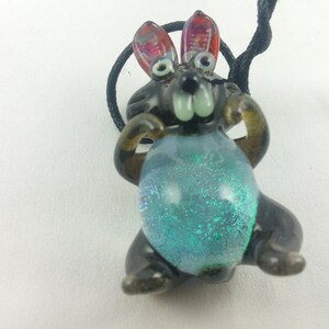 May include: A glass rabbit charm with black and brown details, red ears, and a blue iridescent belly. The charm is hanging from a black cord.
