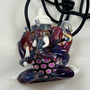 May include: A glass pendant necklace featuring a detailed depiction of the Hindu deity Ganesha. The pendant is made of multi-colored glass with a white crown and intricate details. The pendant is attached to a black cord.