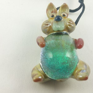 May include: A glass pendant in the shape of a rabbit with a green, glittery belly. The rabbit has brown, orange, and white details and a black cord.
