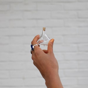 May include: A small, clear glass bottle with a cork stopper. The bottle has a rounded base and a narrow neck. The bottle is held in a hand with gold nail polish and silver rings. The background is a white brick wall.