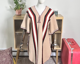 May include: A beige knit poncho with a V-neck and short sleeves, featuring vertical stripes in red, black, and white. The poncho is displayed on a mannequin. The edges are trimmed with red and black detailing.