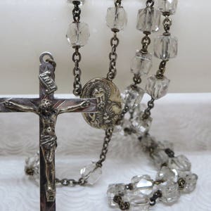 May include: A silver rosary with clear glass beads and a silver crucifix with a figure of Jesus Christ. The crucifix also has a small silver medallion with a figure of a saint.