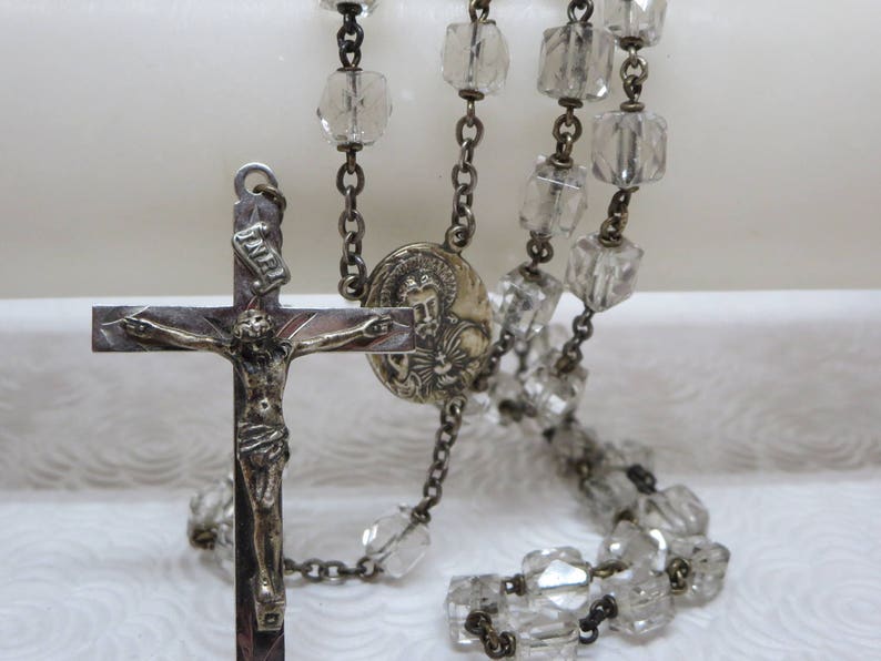 May include: A silver rosary with clear glass beads and a silver crucifix with a figure of Jesus Christ. The crucifix also has a small silver medallion with a figure of a saint.