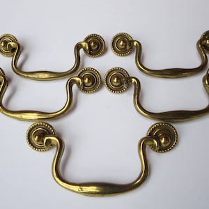 May include: Six antique brass drawer pulls with a decorative design. Each pull has a rounded top and a curved handle.