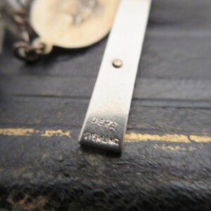 May include: Close-up of a silver cross with the word "DEKA" stamped on the back.