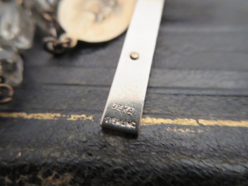 May include: Close-up of a silver cross with the word "DEKA" stamped on the back.