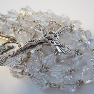 Op de afbeelding: Een zilveren rozenkrans met heldere kristallen kralen en een zilveren kruisbeeld met een figuur van Jezus Christus. Het kruisbeeld heeft een gedetailleerd ontwerp en de rozenkrans is geregen aan een zilveren ketting.
