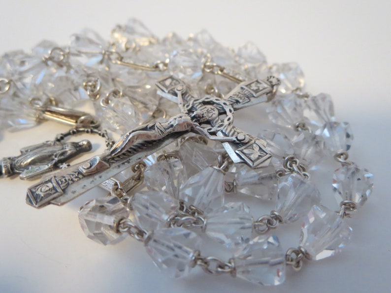 Op de afbeelding: Een zilveren rozenkrans met heldere kristallen kralen en een zilveren kruisbeeld met een figuur van Jezus Christus. Het kruisbeeld heeft een gedetailleerd ontwerp en de rozenkrans is geregen aan een zilveren ketting.
